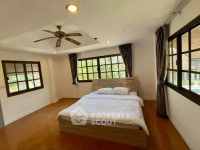 Spacious bedroom with large windows and wooden flooring, featuring a cozy bed and ceiling fan.