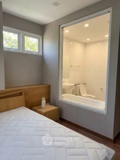Modern bedroom with a unique window view into a sleek bathroom, featuring a minimalist design and natural light.