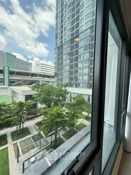 Modern high-rise building view from window with lush green garden and clear blue sky.