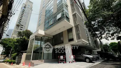 Modern urban building exterior with glass facade and parking area, surrounded by lush greenery.