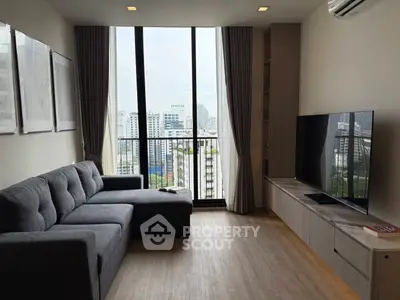 Modern living room with city view, featuring a sleek grey sofa and large flat-screen TV.