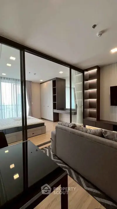 Modern open-plan living room with glass partition to bedroom, featuring sleek furniture and natural light.