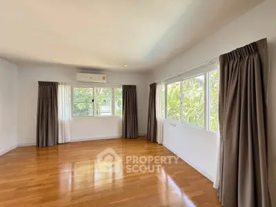 Spacious empty room with hardwood floors and large windows offering lush garden views.