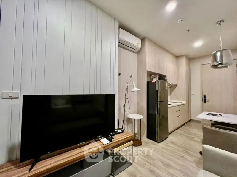 Modern living room with sleek TV, stylish furniture, and compact kitchen in open layout apartment.