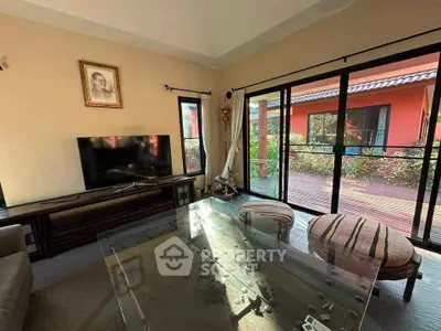 Spacious living room with glass table and large TV, opening to a sunny patio.
