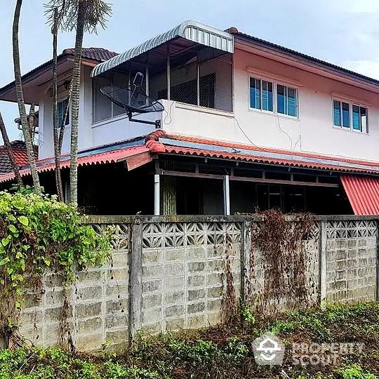 Charming two-story home with a spacious balcony and satellite dish, surrounded by lush greenery and a sturdy concrete fence for privacy.