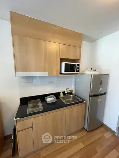 Modern compact kitchen with wooden cabinets, microwave, and fridge in a cozy apartment.