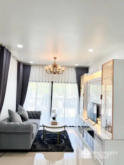 Elegant living room with plush grey sofa, chic glass coffee table, and a classic chandelier, complemented by stylish drapes and a modern entertainment unit.