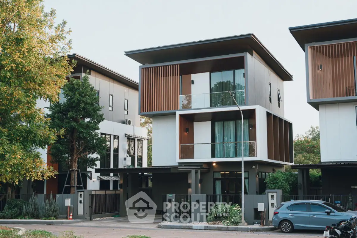 Modern three-story house with sleek design and large windows, surrounded by greenery and parked car.