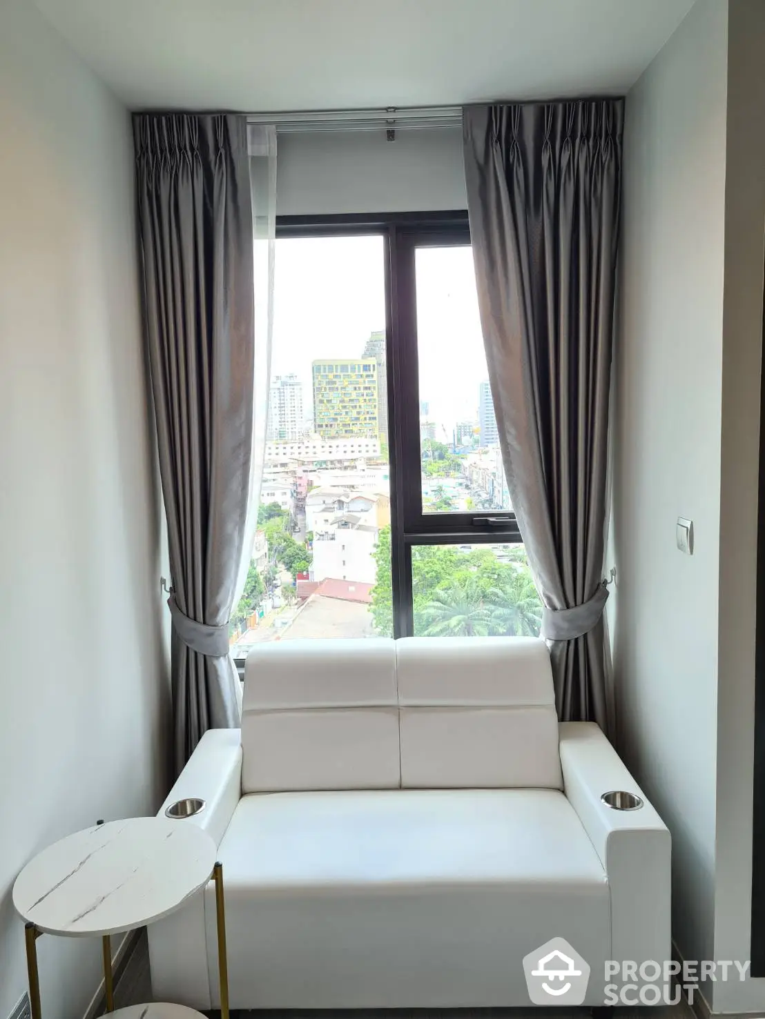 Chic urban living space with a plush white sofa by a large window offering a scenic city view, complemented by elegant grey curtains.