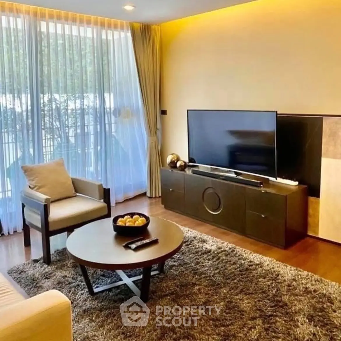 Modern living room with large TV, cozy seating, and natural light through floor-to-ceiling windows.