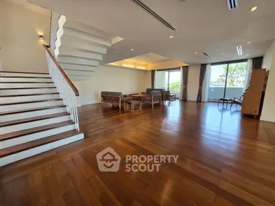 Spacious living room with wooden flooring and modern furniture, featuring large windows and a staircase.