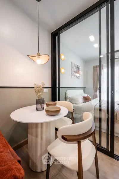 Modern dining area with stylish furniture and view into cozy bedroom through glass partition.