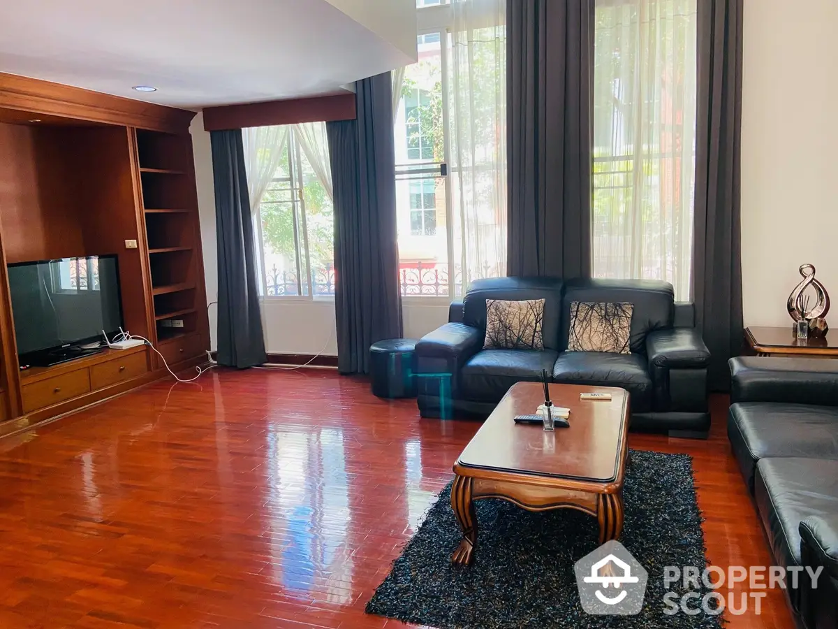 Spacious living room with gleaming hardwood floors, large windows allowing ample natural light, and a comfortable black leather sofa set, perfect for entertaining guests or relaxing.