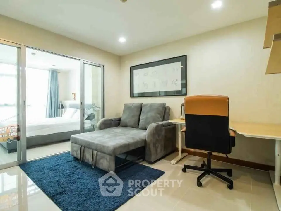 Modern living room with cozy sofa and study area, adjacent to a bright bedroom with glass sliding doors.