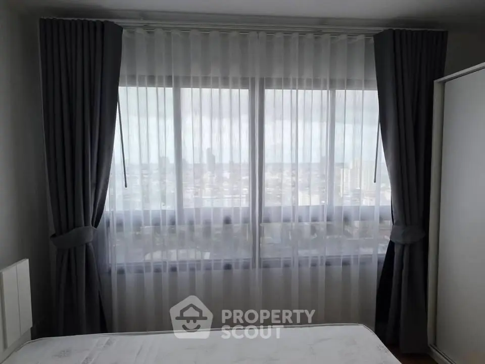 Bright bedroom with large window and city view, featuring elegant curtains and a cozy atmosphere.