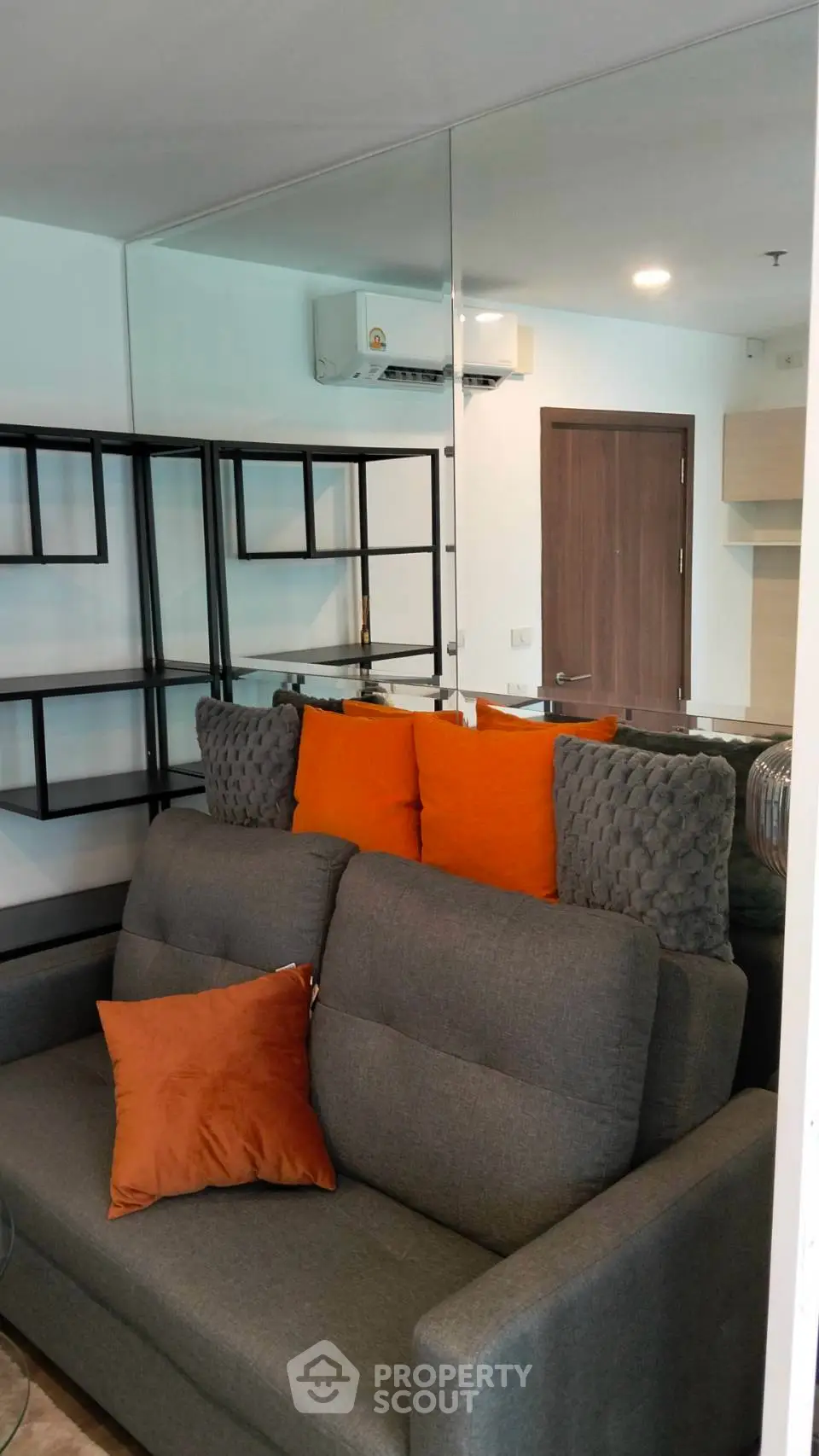 Modern living room with stylish gray sofa and vibrant orange cushions, featuring sleek shelving and a mirrored wall.