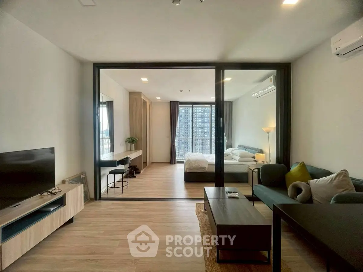 Modern open-plan living room with glass partition leading to a cozy bedroom, featuring stylish furniture and city views.