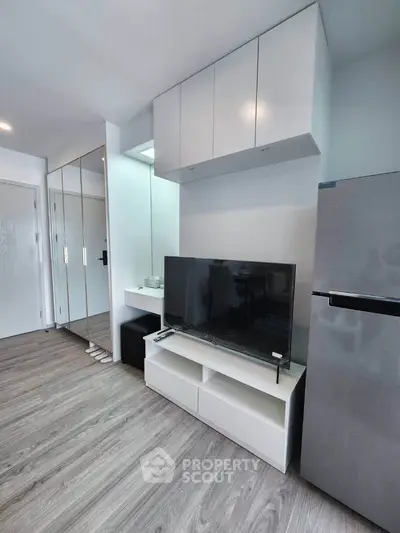 Modern living room with sleek TV stand, mirrored closet, and stainless steel fridge in a compact apartment.