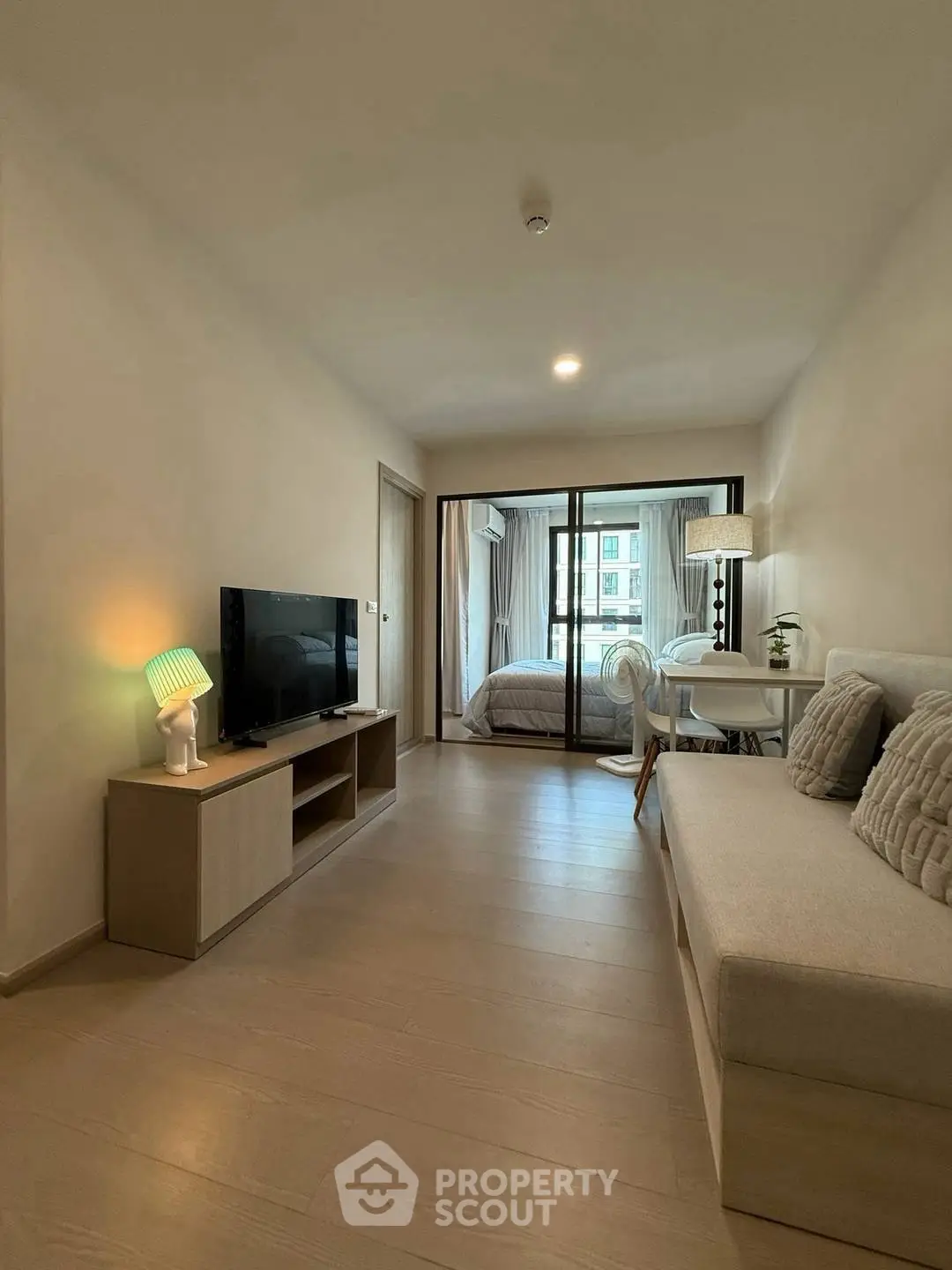 Modern living room with cozy sofa, TV, and view into bedroom through glass doors.