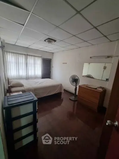 Cozy bedroom with natural light, featuring a bed, fan, and dresser, ideal for restful living.