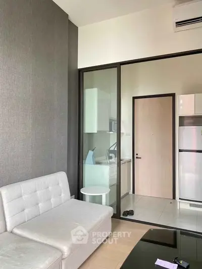 Modern living room with sleek sofa and glass partition leading to kitchen area.