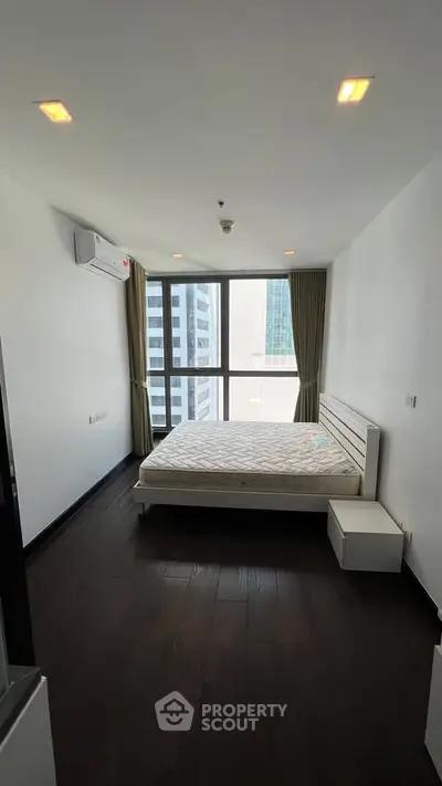 Modern bedroom with large window and city view, featuring a minimalist design and dark wood flooring.