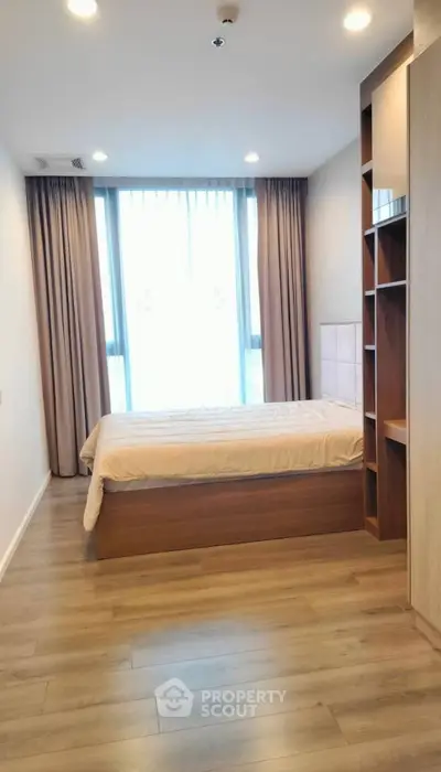Modern bedroom with wooden flooring and large window, featuring built-in shelves and cozy ambiance.