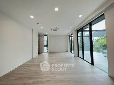Spacious modern living room with large windows and balcony access, featuring sleek wooden flooring and recessed lighting.