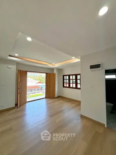 Spacious room with wooden flooring and large windows opening to a balcony.