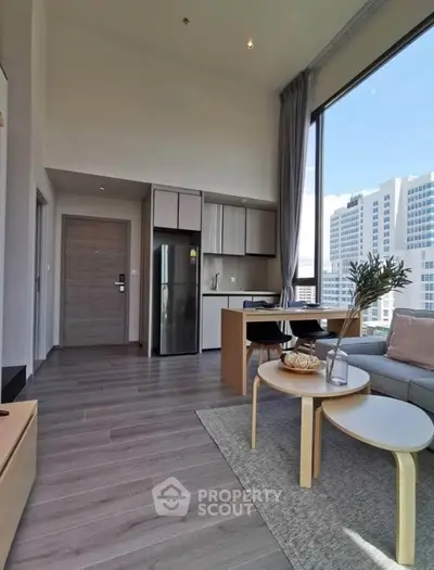 Modern living room with open kitchen and city view, featuring sleek furniture and large windows.