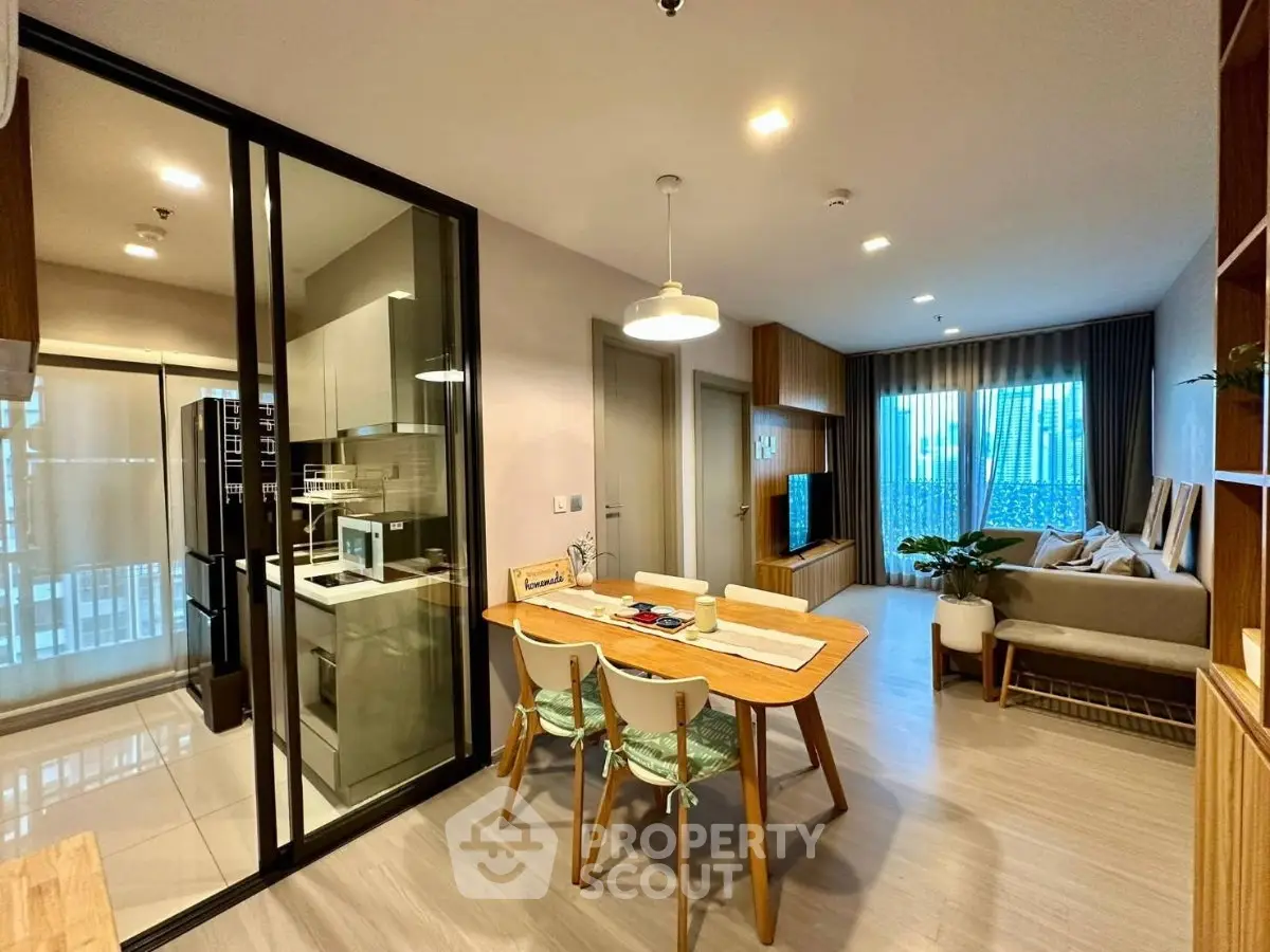 Modern open-plan living room with dining area and glass-enclosed kitchen, featuring stylish decor and natural light.
