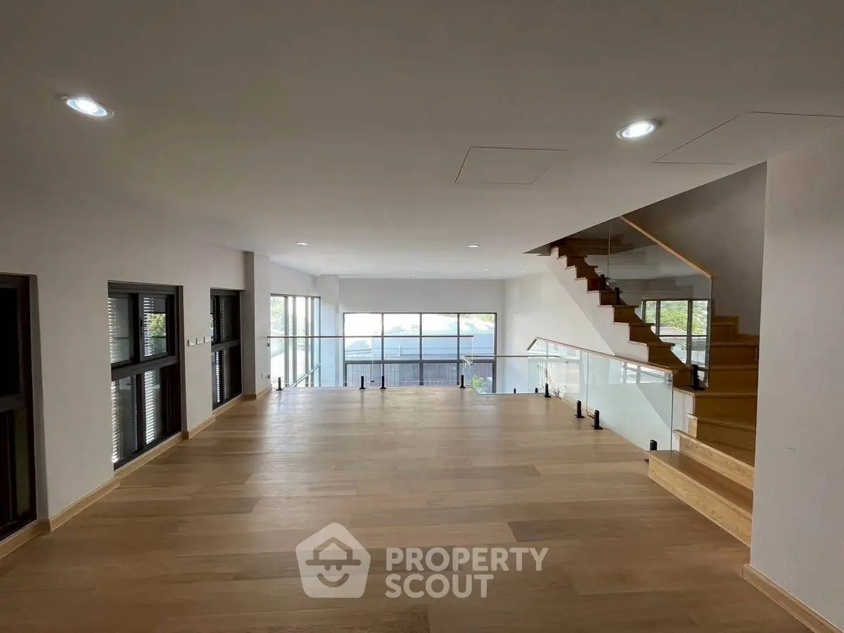 Spacious modern living room with wooden flooring and glass staircase, offering ample natural light.