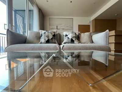 Modern living room with stylish sofa and glass coffee table, featuring geometric cushions and wooden flooring.