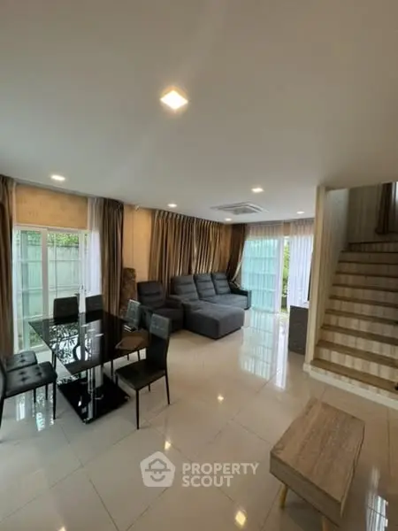 Spacious modern living room with elegant dining area and staircase, featuring large windows and stylish decor.