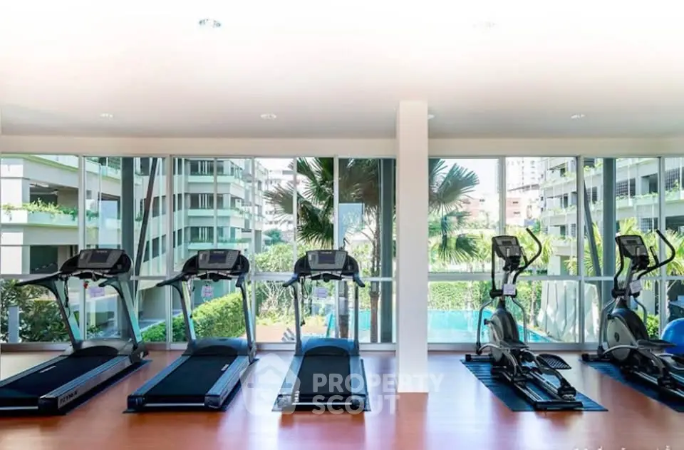 Modern gym with treadmills and exercise bikes overlooking lush greenery and pool view.