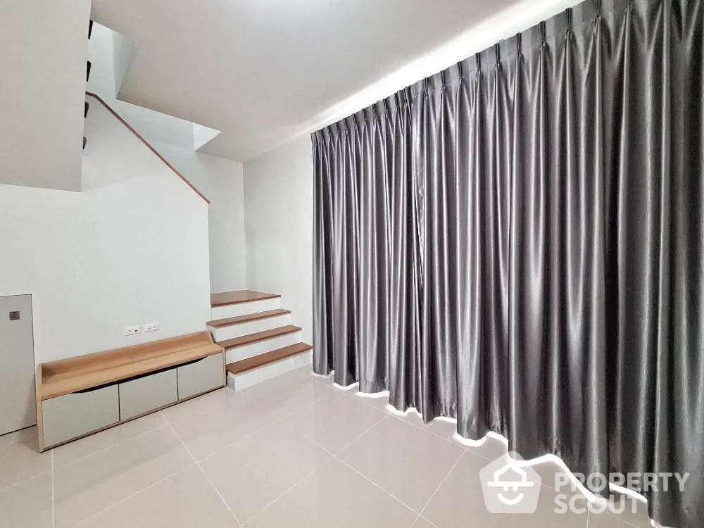 Spacious and modern living room with elegant dark curtains and clean white tiled flooring, featuring a unique staircase leading to an upper level.