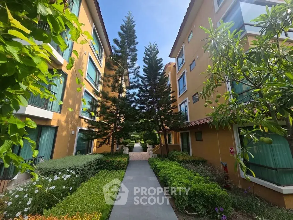 Charming garden pathway between two residential buildings with lush greenery and tall trees.
