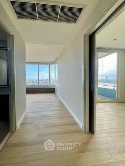 Spacious hallway with wooden flooring leading to a bright room with large windows and balcony view.