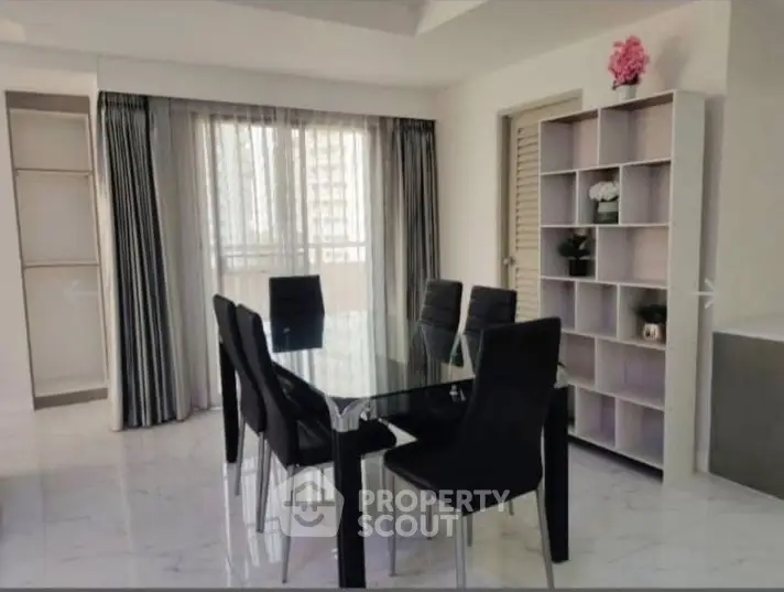 Elegant dining area with glass table and modern chairs, featuring a stylish bookshelf and large window for natural light.