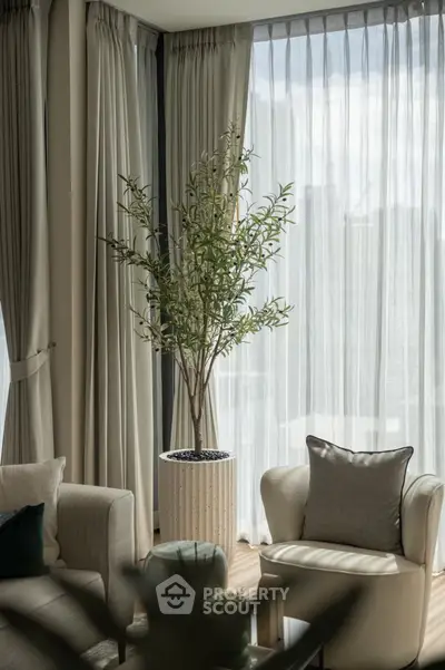 Elegant living room with natural light, featuring cozy seating and a stylish potted plant by large windows.