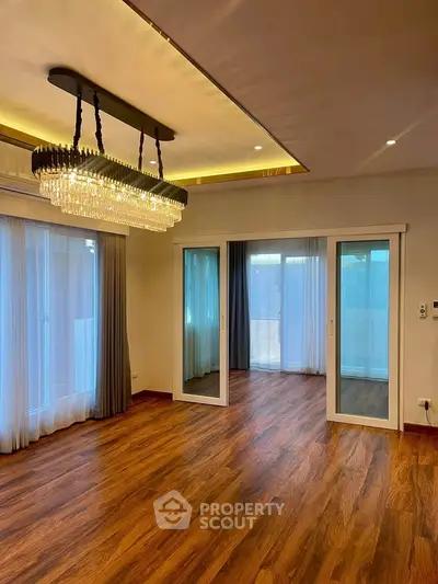 Spacious living room with elegant chandelier and wooden flooring, featuring large windows for natural light.