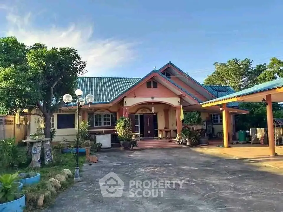 Charming single-story home with blue roof and spacious driveway, surrounded by lush greenery.