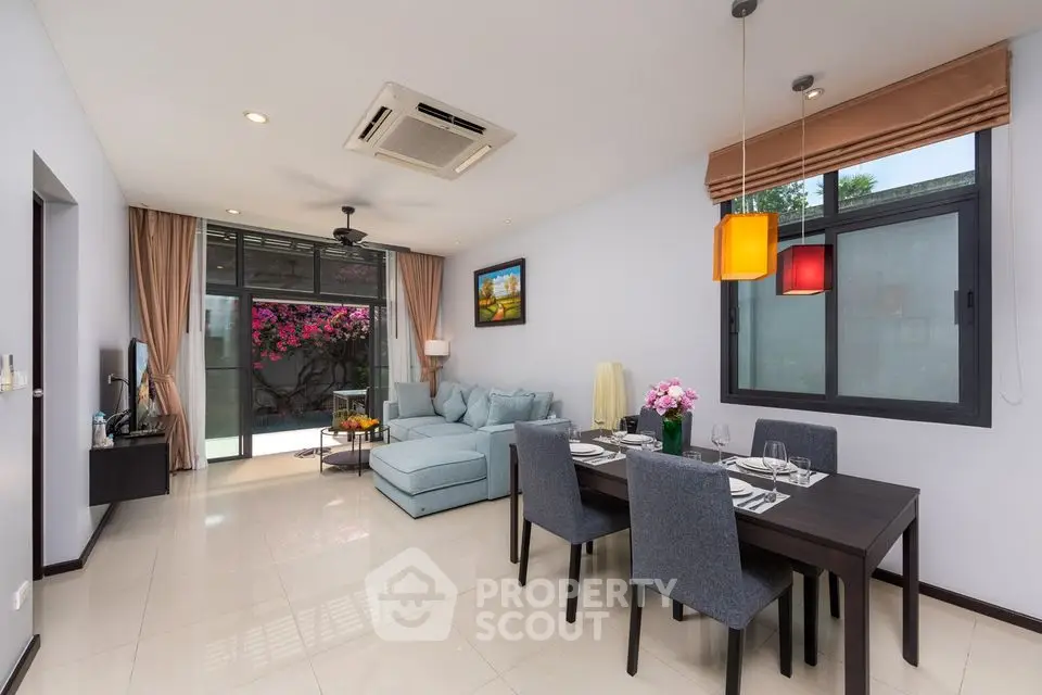 Spacious and modern living room with sleek dining set, comfortable sofa, and vibrant hanging lights, leading to a cozy balcony with pink flowers.
