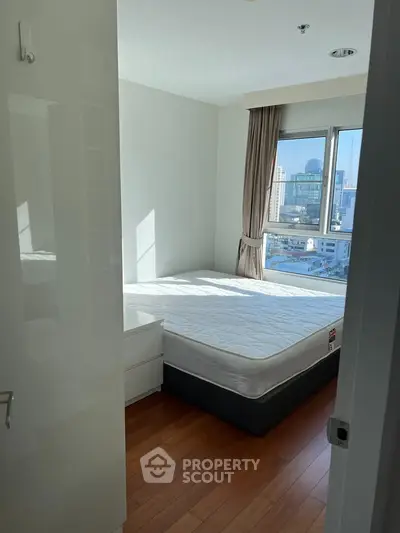 Bright bedroom with large window and city view, featuring a comfortable bed and wooden flooring.