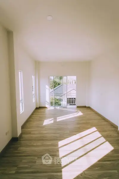 Spacious sunlit room with wooden flooring and large windows offering a serene view.