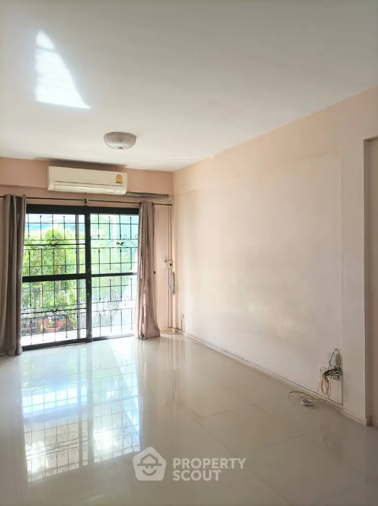 Spacious and bright living room with glossy tiled flooring, large windows leading to a balcony with a serene view, and an air conditioning unit for comfort.