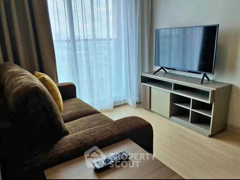 Modern living room with cozy sofa and flat-screen TV, featuring natural light through sheer curtains.