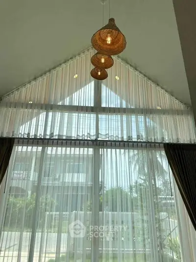 Elegant living room with high ceiling and large windows, featuring stylish pendant lights and sheer curtains.