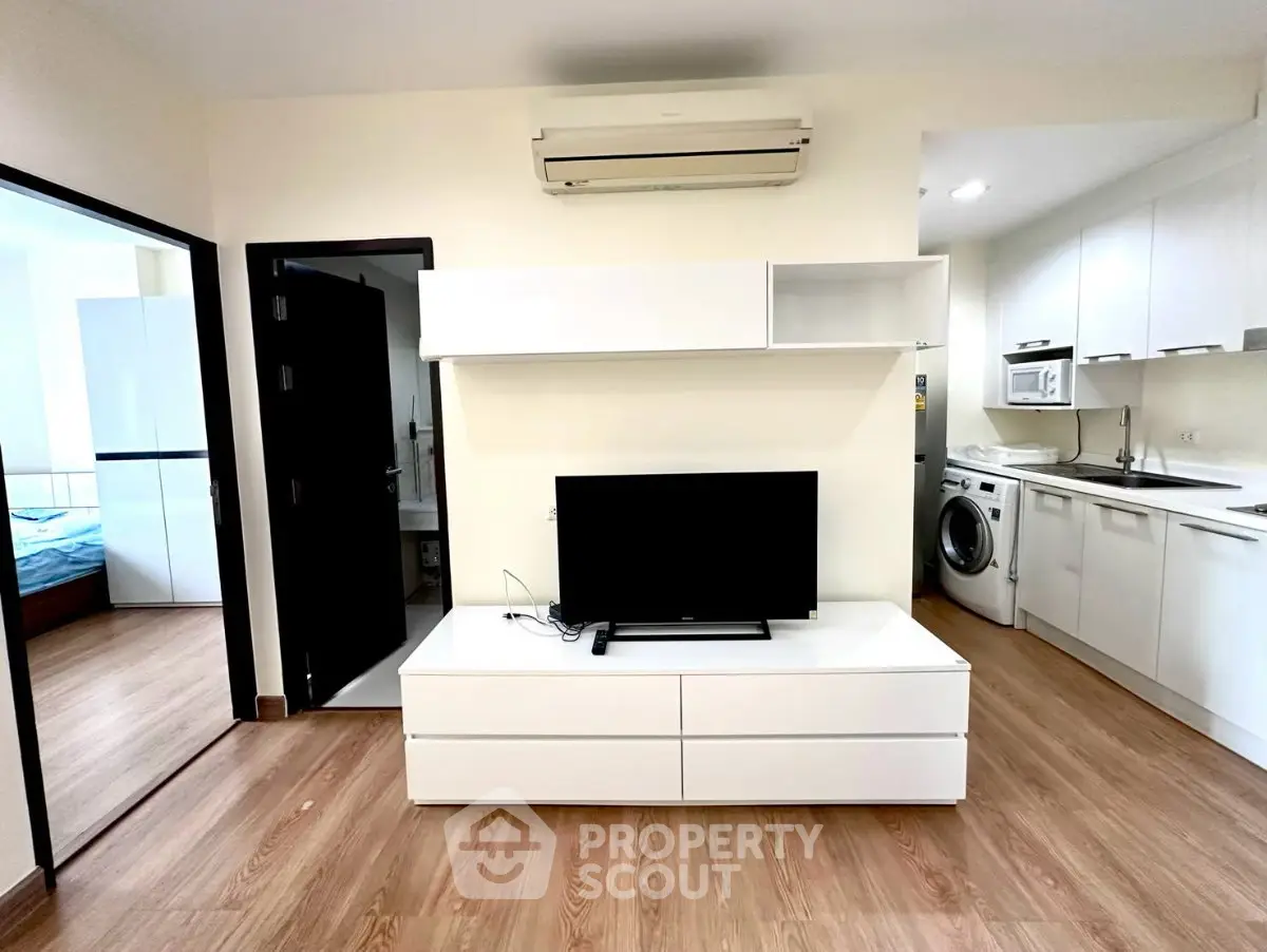 Modern apartment interior with open kitchen, living area, and bedroom view, featuring sleek white furniture and wood flooring.